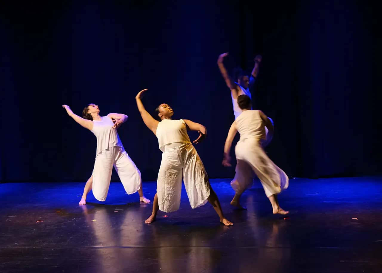 Dancers in white leaning back with their right arms raised on a dark stage.